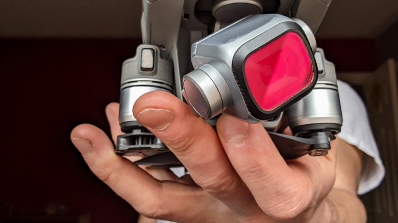 A hand holding a compact drone with a red-tinted ND filter attached to the gimbal-mounted camera lens.