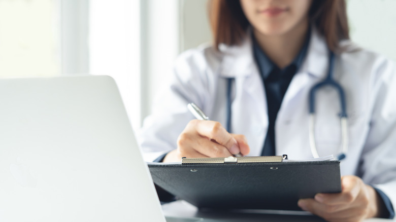 A doctor with a clipboard sitting behind a laptop in a clinical setting.