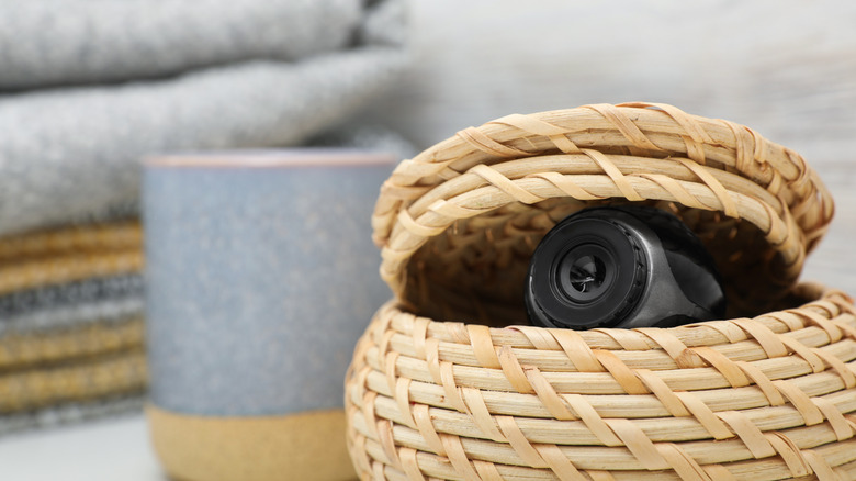 A compact security camera lens peeking through the weave of a wicker storage basket.