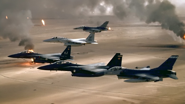 A formation of F-16 fighter jets flying over a desert landscape with burning oil wells in the background.