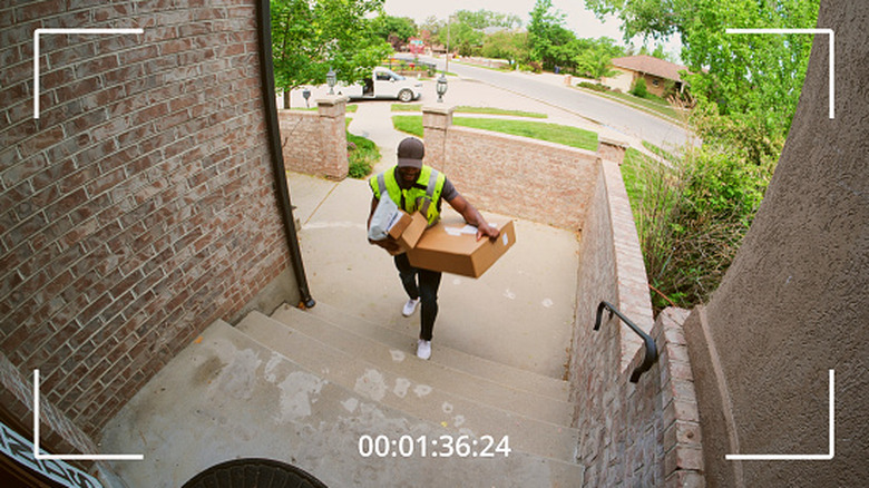 A fish-eye lens perspective from a doorbell camera showing a delivery worker placing a cardboard package on a doorstep.