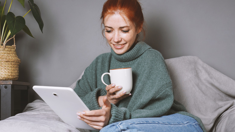 A woman comfortably holding a small tablet while sitting with a cup of coffee.