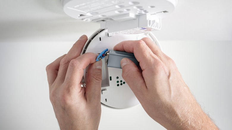 A person's hands installing a fresh battery into a smoke detector unit.